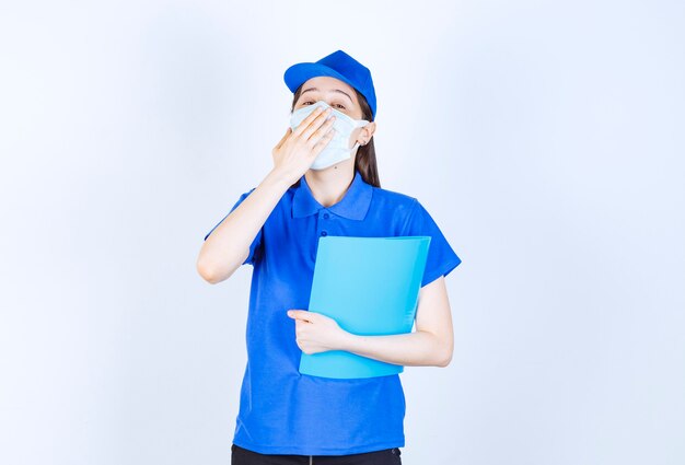 Foto de mujer joven en máscara médica sosteniendo una carpeta y bostezando.