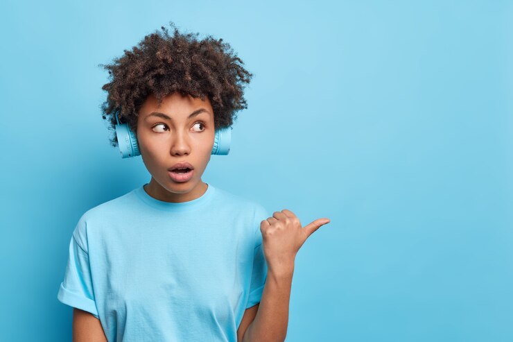 Foto gratuito la foto de una mujer joven de pelo rizado conmocionada apunta hacia un espacio en blanco sorprendido al notar la increíble oferta de venta escucha la pista de audio en auriculares vestidos casualmente aislados sobre una pared azul