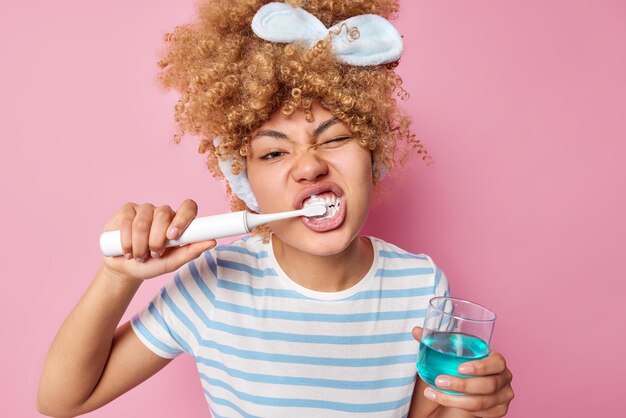 Foto de una mujer joven con el pelo rizado y peinado, cepilla los dientes, usa un cepillo eléctrico, se somete a un procedimiento de blanqueamiento, sostiene un vaso de enjuague bucal, usa una camiseta casual a rayas aislada sobre un fondo rosado