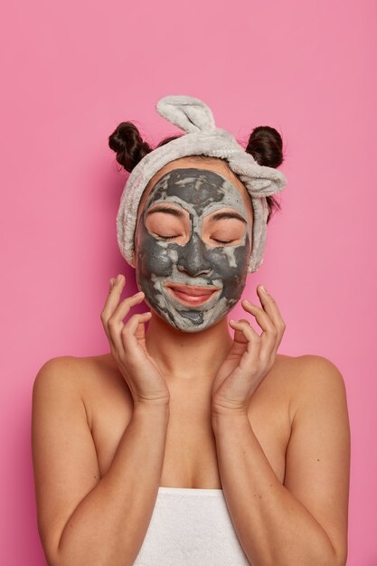 Foto de mujer joven que se relaja con mascarilla facial, toca su piel fresca, disfruta de tratamientos de belleza, se para envuelta en una toalla, cierra los ojos, modela contra la pared rosada, se preocupa por la tez.