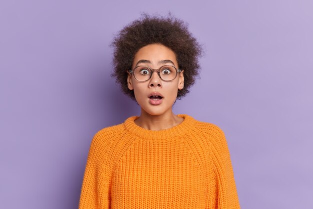 La foto de una mujer joven rizada sorprendida mira fijamente los ojos saltones mantiene la boca abierta tiene expresión de sorpresa usa lentes ópticos suéter de punto naranja.