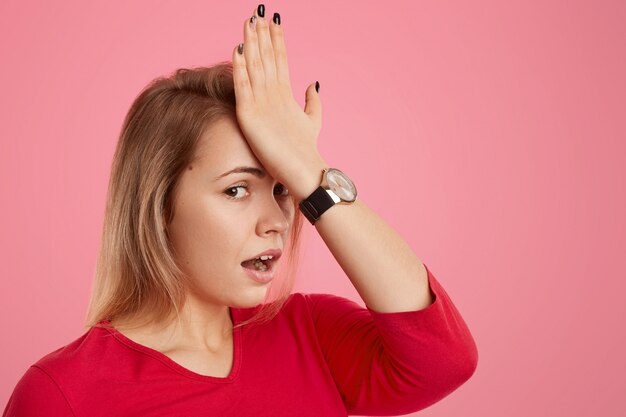 Foto de mujer joven rubia frustrada con piel sana, mantiene la mano en la frente