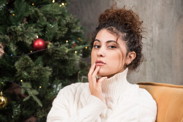 Foto de mujer joven sentada en una cómoda silla cerca del árbol de Navidad