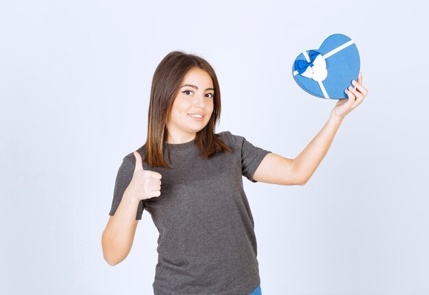 Foto de mujer joven sonriente mostrando un pulgar hacia arriba y sosteniendo una caja de regalo en forma de corazón.