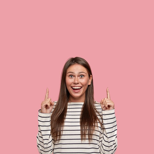 Foto de mujer llena de alegría con una sonrisa radiante, apunta con el dedo índice hacia arriba