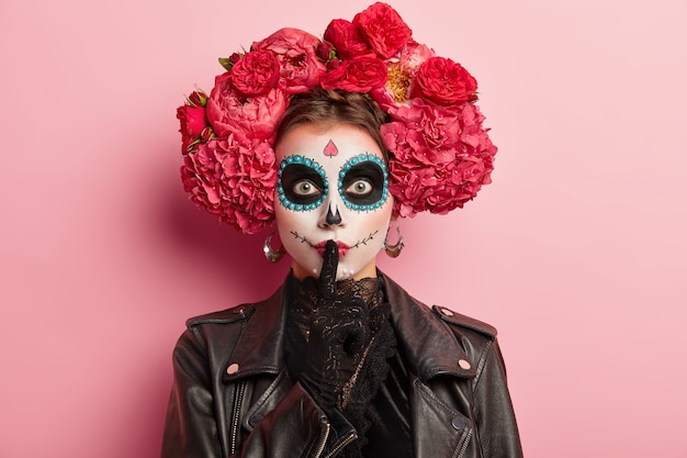 Foto de mujer con maquillaje tradicional y flores en el cabello, hace gesto de silencio, mantiene el dedo índice sobre los labios pintados, se prepara para la horrible fiesta de la muerte, vestida con un traje negro, aislado en rosa