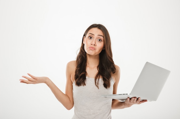 Foto de mujer molesta con largo cabello castaño levantando las manos y expresando confusión mientras sostiene la computadora personal plateada, aislada sobre la pared blanca