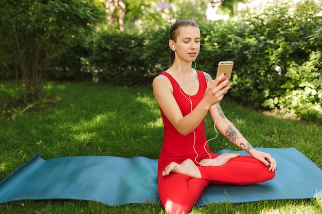 Foto de una mujer con mono rojo sentada en una pose de loto en una alfombra de yoga en el parque. Señorita en auriculares usando teléfono celular mientras practica yoga al aire libre