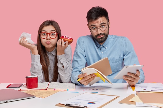 Foto gratuita foto de mujer morena divertida que come una deliciosa rosquilla, tiene la boca llena mientras su pareja se da cuenta de noticias impactantes, mira el bloc de notas, sostiene el panel táctil