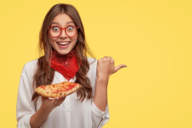 Foto de una mujer morena sorprendida y emotiva con una gran sonrisa, viste un pañuelo rojo, sostiene una rebanada de pizza, señala con el pulgar a un lado, modelos contra la pared amarilla para su contenido publicitario. Plato sabroso