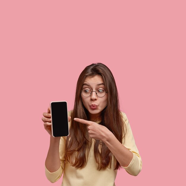 La foto de una mujer morena sorprendida mantiene los labios doblados, indica con el dedo índice en la pantalla simulada del celular, demuestra algo asombroso