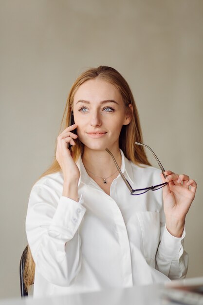 Foto de una mujer de negocios rubia joven alegre en la oficina en el interior trabaja con ordenador portátil y teléfono móvil.