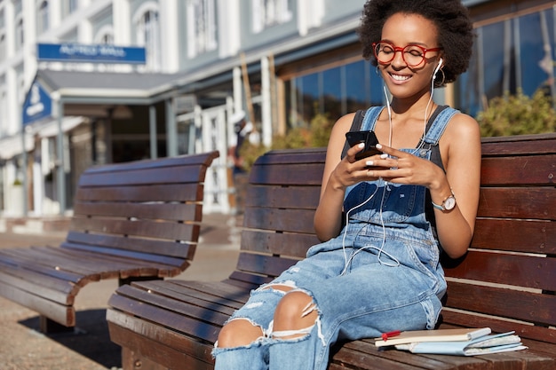 Foto de mujer negra alegre sostiene celular, escribe mensajes de texto, usa audífonos, escucha música, vestida con overoles andrajosos, modela al aire libre, disfruta de la lista de reproducción. Tecnologías modernas, comunicación online