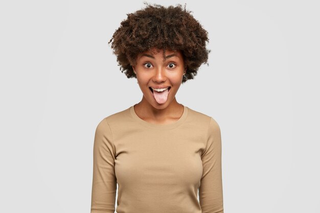Foto de mujer negra divertida muestra lengua, tiene expresión juguetona, peinado afro