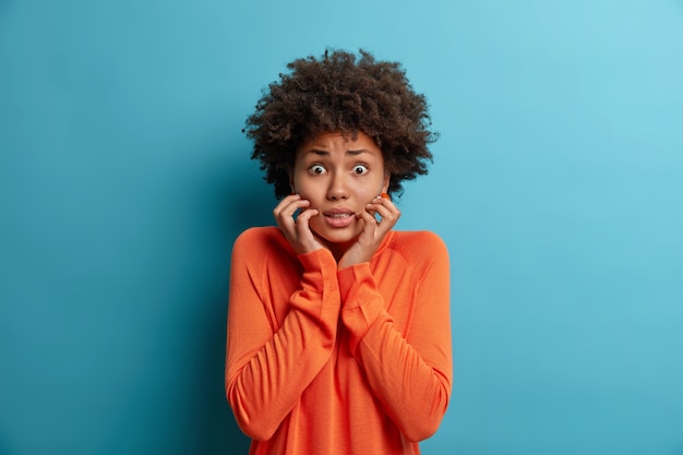 Foto de mujer nerviosa asustada agarra la cara y mira con expresión preocupada, ve fobia, tiene miedo de hablar, usa un jersey naranja, aislado en una pared azul. Concepto de reacción humana