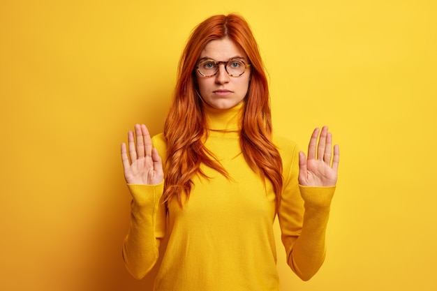 Foto de mujer pelirroja joven seria muestra gesto de parada hace señal prohibida tira de las palmas hacia, lleva gafas ópticas y cuello alto.