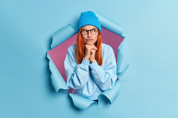 Foto de mujer pelirroja seria sostiene las manos debajo de la barbilla parece disgustado a un lado sonríe con insatisfacción usa un jersey azul y un sombrero se rompe a través del agujero de papel