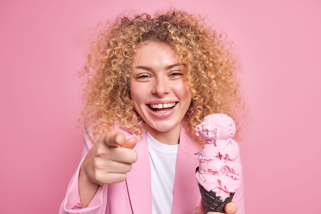 La foto de la mujer de pelo rizado llena de alegría se ríe alegremente apunta directamente sostiene apetitosas sonrisas de helado muestra ampliamente los dientes blancos aislados sobre la pared rosa. Concepto de alimentación poco saludable