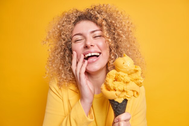 Foto de mujer de pelo rizado llena de alegría se siente divertida sonríe ampliamente mantiene los ojos cerrados disfruta comiendo deliciosos helados de sabor a mango posa con delicioso postre helado aislado en la pared amarilla.