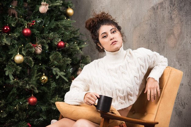 Foto de mujer pensativa sentada en una silla cómoda con una taza de bebida