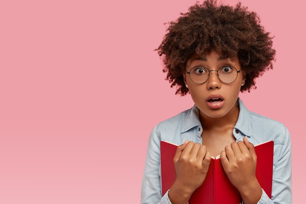 Foto de una mujer de piel oscura asustada sorprendida que lleva un libro de texto rojo, lee algo asombroso, jadea de miedo, vestida con ropa de mezclilla, modelos sobre una pared rosada con espacio libre para su anuncio