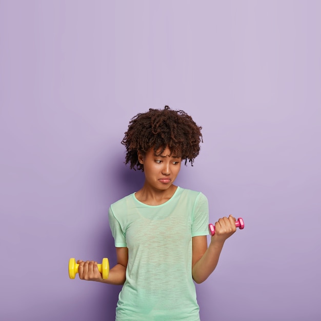 Foto de mujer de piel oscura insatisfecha que levanta pesas, hace ejercicio, quiere ser fuerte y saludable, vestida con una camiseta informal, levanta mucho peso