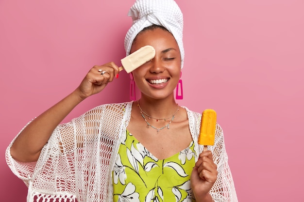 La foto de una mujer de piel oscura positiva se divierte y sostiene un delicioso helado frío, cubre los ojos con una paleta, tiene una amplia sonrisa, vestida con ropa doméstica, aislada en la pared rosa. Verano, alegría, comiendo