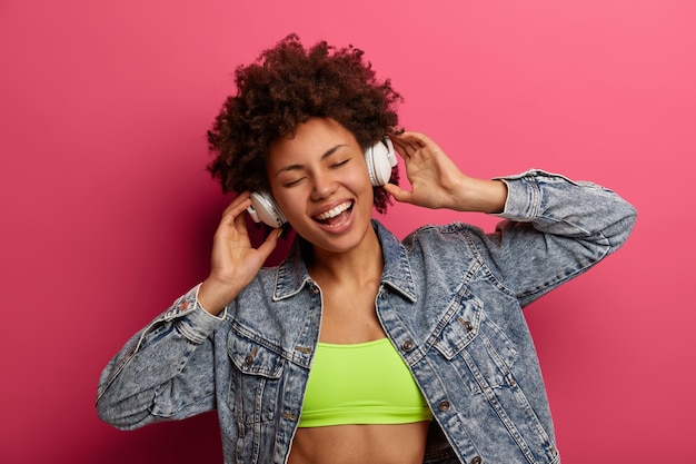 Foto gratuita foto de mujer de piel oscura que escucha música en auriculares, sonríe alegremente, inclina la cabeza y se viste con ropa a la moda, se siente complacida de escuchar su canción favorita, baila contra la pared rosada