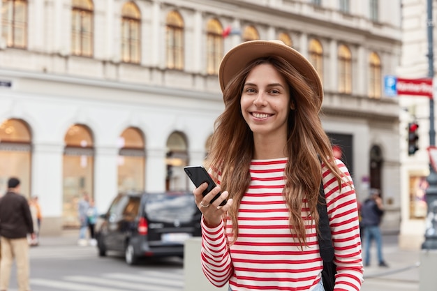 Foto de mujer positiva usa teléfono celular para navegar en la ciudad, verifica notificación