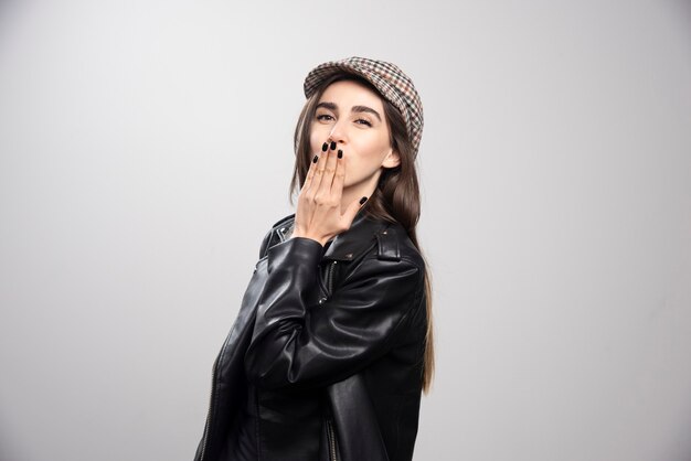 Foto de una mujer que sopla un beso al aire en chaqueta de cuero negro y gorra.