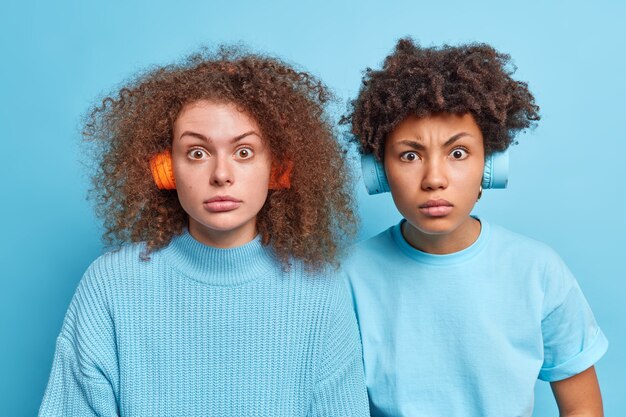 Foto de mujer de raza mixta grave sorprendida mirada perpleja escuchar música a través de auriculares inalámbricos vestidos casualmente aislado sobre la pared azul. Concepto de ocio de estilo de vida de amistad