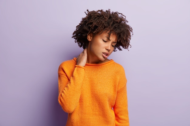 Foto gratuita foto de mujer rizada descontenta que lleva la mano en el cuello, sufre de dolores terribles, trabaja duro, tiene un estilo de vida sedentario, cabello espeso y rizado oscuro, usa jersey naranja, modelos sobre pared violeta.