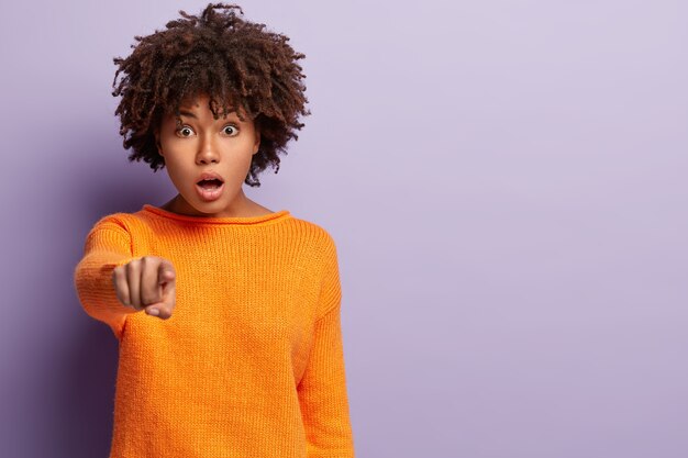 La foto de una mujer rizada emocional sorprendida sobresaltada señala con el dedo índice recto, abre la boca por la conmoción, demuestra algo al frente, usa ropa informal, modelos sobre una pared púrpura