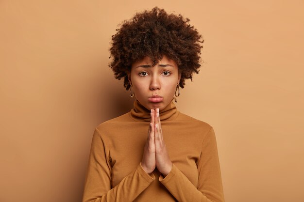 La foto de una mujer rizada de piel oscura y sombría pide lo mejor, mantiene las palmas juntas, pide perdón, se viste informalmente, se aísla sobre una pared marrón, pide. Por favor hazme un favor