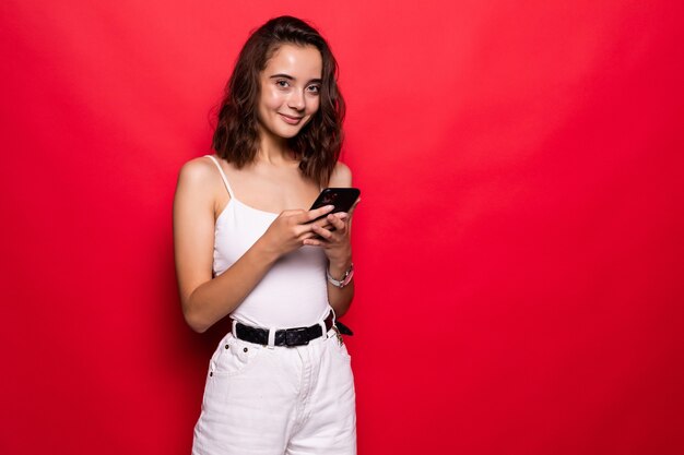 Foto de mujer satisfecha mirando a la cámara mientras usa el teléfono celular con alegría sobre rojo