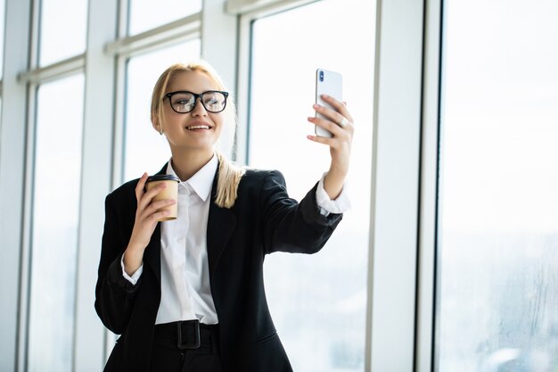 Foto de mujer secretaria en ropa formal de pie sosteniendo café para llevar en la mano y tomando selfie en teléfono móvil en la oficina