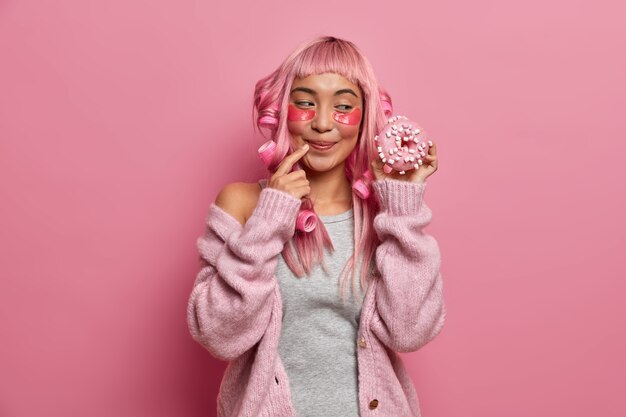 Foto de mujer sonriente con aspecto de goog es golosa y se ve con apetito en una deliciosa rosquilla, usa rizadores de pelo, tiene peinado rosa