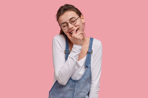 Foto de mujer sonriente complacida cubre la boca, cierra los ojos de placer, se ríe de una buena broma, vestida con ropa de moda para jóvenes