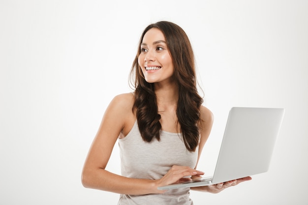 Foto de mujer sonriente con largo cabello castaño mirando a otro lado mientras trabajaba en la computadora personal plateada, aislada sobre la pared blanca