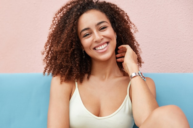 Foto de mujer sonriente positiva con amplia sonrisa encantadora vestida de manera informal y recrea en casa, se siente relajada y cómoda