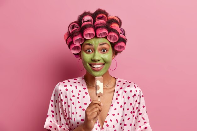 La foto de una mujer sonriente sonríe ampliamente, se aplica una mascarilla humectante verde, come un delicioso helado en un palito, usa rulos, una bata de seda, disfruta del verano. Ama de casa con postre helado