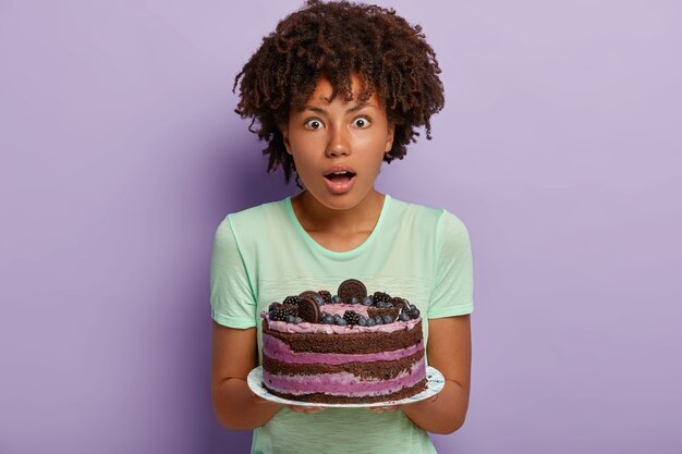 Foto de mujer sorprendida de piel oscura con corte de pelo rizado, sostiene un delicioso pastel, los invitados sorprendidos ya comieron en el umbral, vestidos con ropa casual, postre sabroso horneado