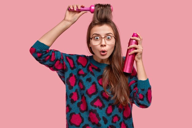 Foto de mujer sorprendida que se peina, rocía flecos, usa cepillo para el cabello, usa anteojos y suéter