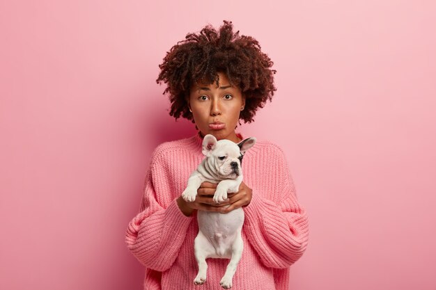 Foto de mujer triste y deprimida que se siente infeliz porque su mascota está enferma, lleva un perro pequeño al veterinario, frunce los labios, pide curar al animal, usa un jersey rosa suelto, posa junto con el cachorro en el interior sobre una pared rosada