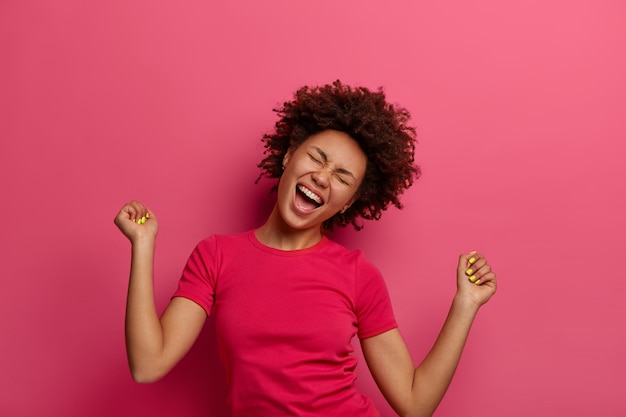 Foto de mujer triunfante llena de alegría que golpea el puño, inclina la cabeza y se ríe de alegría, celebra su propio éxito, usa una camiseta informal, obtiene la victoria y logra la meta, posa sobre una pared rosa.