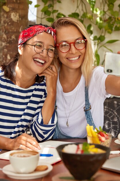 Foto de mujeres alegres de raza mixta cooperan juntas, hacen una foto selfie para compartir en las redes sociales