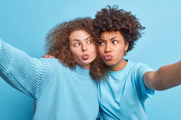 Foto gratuita foto de mujeres divertidas de raza mixta que hacen pucheros con los labios y hacen muecas, abrazan los brazos estirados para hacer selfie, usan ropa casual azul en un tono con juegos de pared y pasan tiempo libre juntos