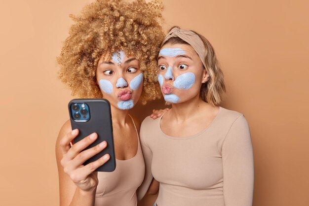 Foto de mujeres jóvenes sorprendidas que aplican máscaras de belleza en la cara hacen muecas en la cámara del teléfono inteligente toman selfie vestidas con ropa informal aisladas sobre fondo beige Amigos y concepto de cuidado de la piel