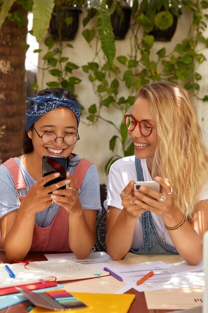 Foto de mujeres de raza mixta complacidas hacen un proyecto juntas, adictas a las tecnologías modernas, intercambian fotos