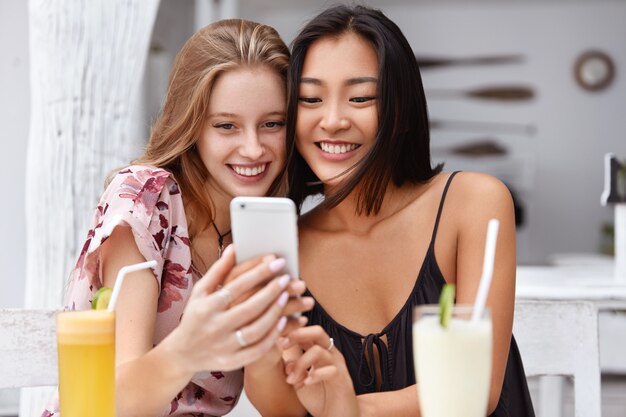 Foto de mujeres de raza mixta complacidas reciben buenas noticias en el teléfono móvil, reciben correos electrónicos o se hacen selfies con teléfonos inteligentes, beben cócteles frescos en la cafetería.